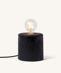 Vintage-style Edison light bulb with visible glowing filaments, mounted on a round charred or dark wooden base with a textured, cracked surface.