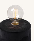 Vintage-style Edison light bulb with visible glowing filaments, mounted on a round charred or dark wooden base with a textured, cracked surface.