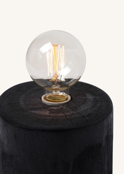 Vintage-style Edison light bulb with visible glowing filaments, mounted on a round charred or dark wooden base with a textured, cracked surface.