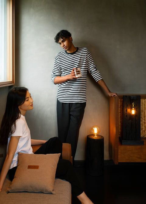 Interior room scene with vintage Edison bulb lamps, showing two people in a casual setting - one seated on a tan couch with a decorative pillow, wearing a white t-shirt, and another standing wearing a black and white striped shirt, with warm ambient lighting creating a cozy atmosphere.